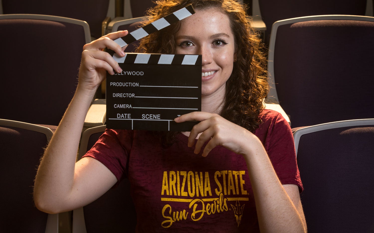 A woman wearing an ASU shirt smiles at the camera while holding a film clapperboard / Credit ASU Online