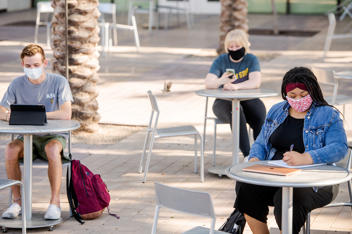 Three people sit outside at separate tables, wearing masks and working on homework or using their phones.