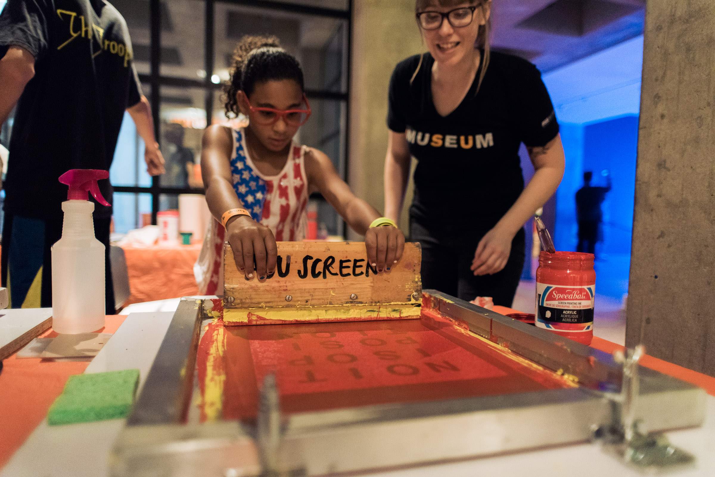Image shows a young girl learning how to make a screenprint under the guidance of an ASU student