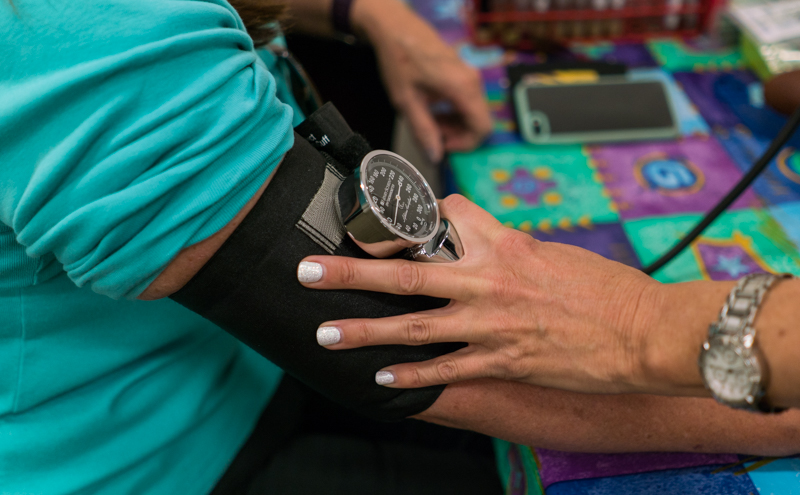 Image of a person's arm with a blood pressure cuff and gauge and another person's hand holding the cuff.