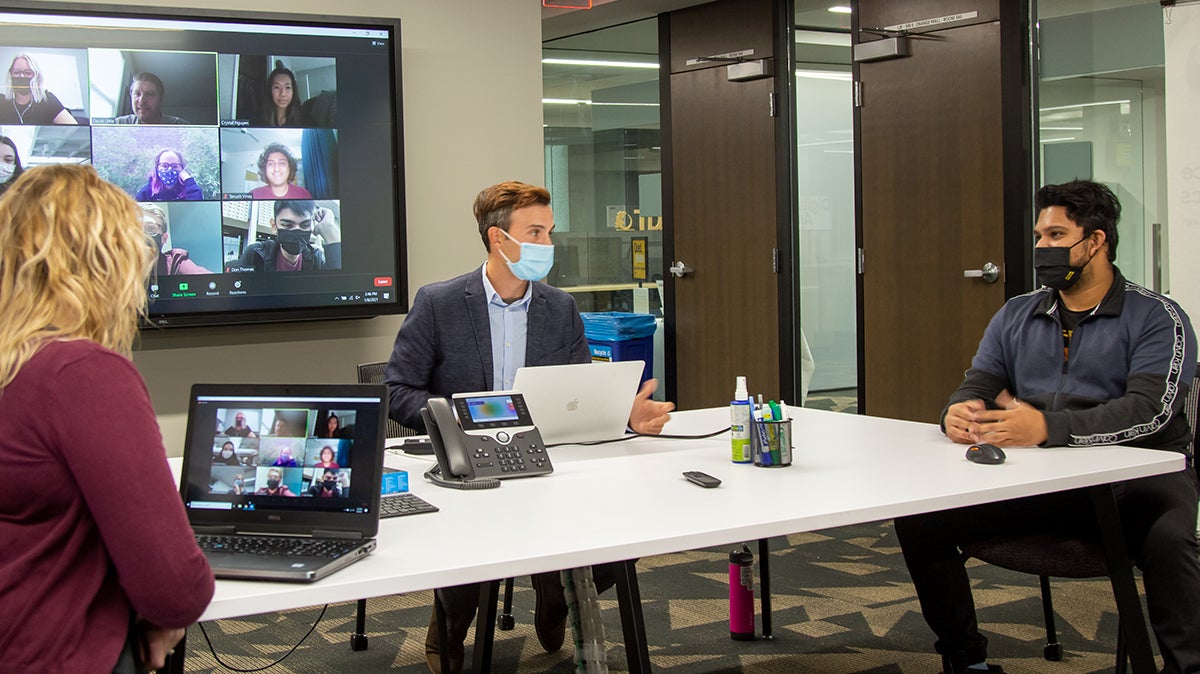 Three people of different genders and ethnicities discussing around a table while interactng with online Zoom attendees