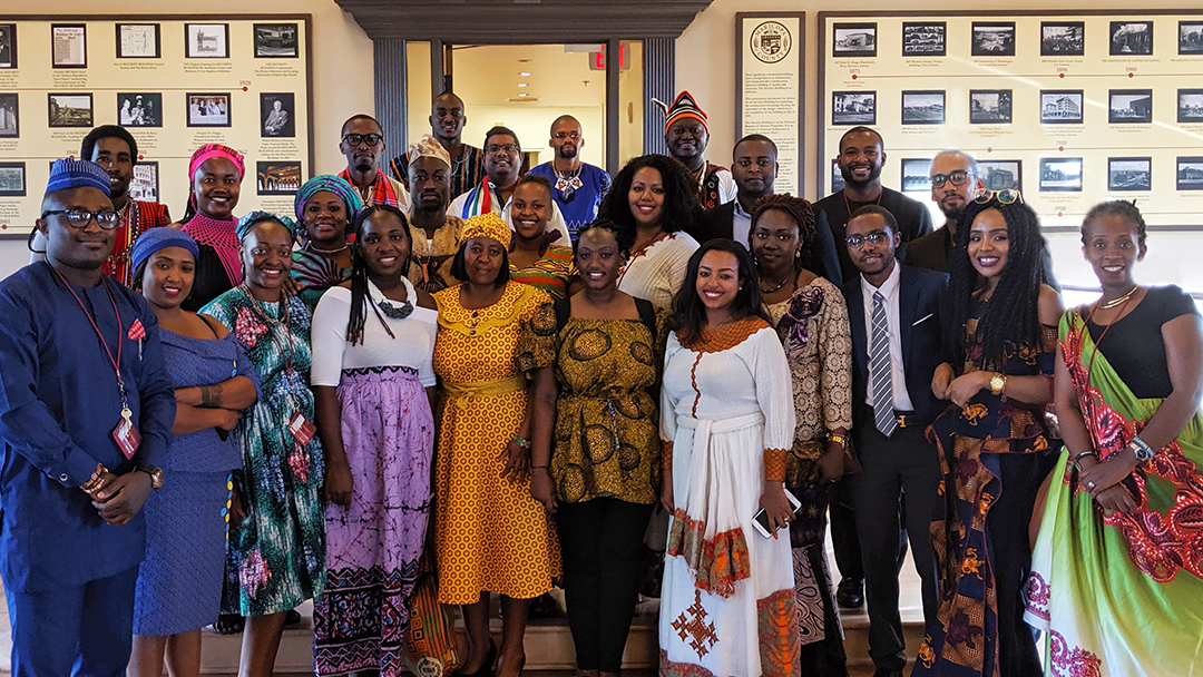 Group of 25 young professionals from Western Africa, wearing traditional dress from their respective countries, standing on stairs in historical room in Maricopa Security building