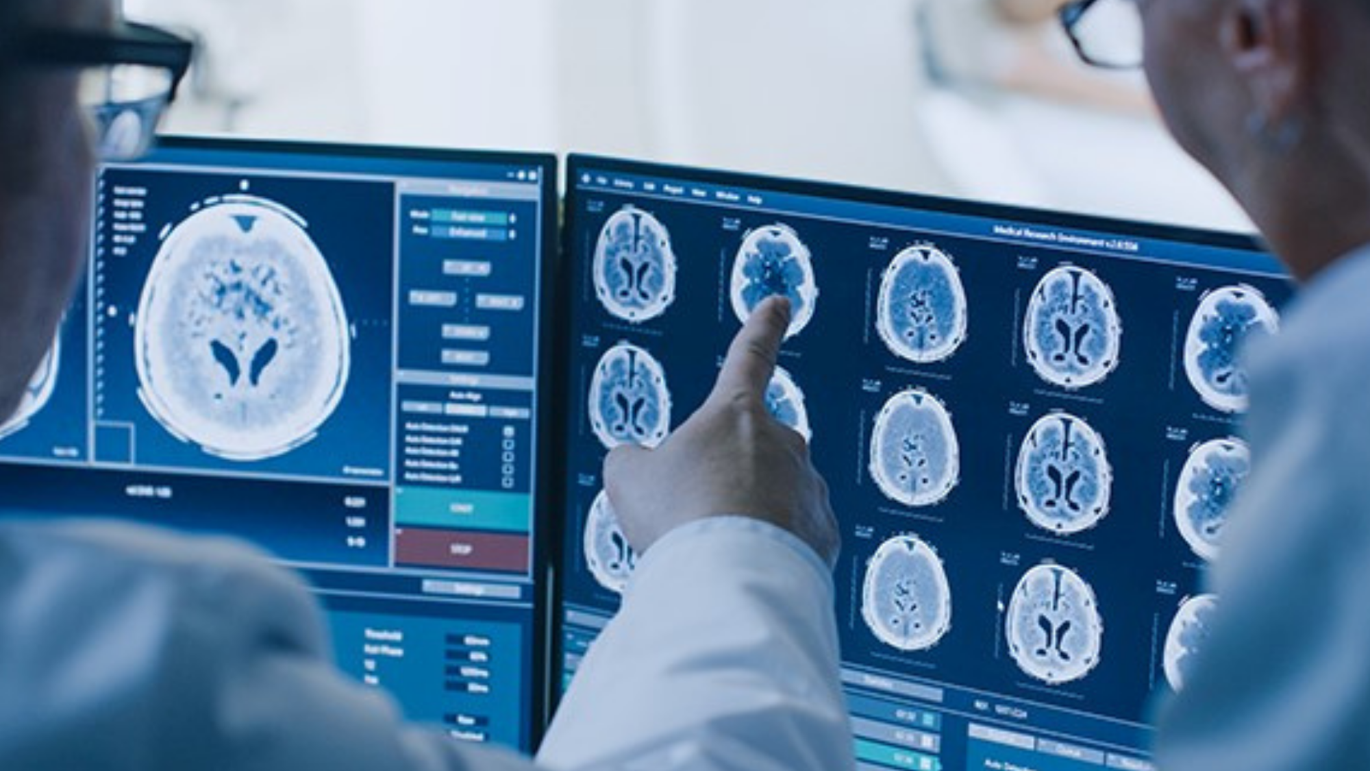 A man wearing glasses points to a computer monitor showing images of brain scans while another man looks on.