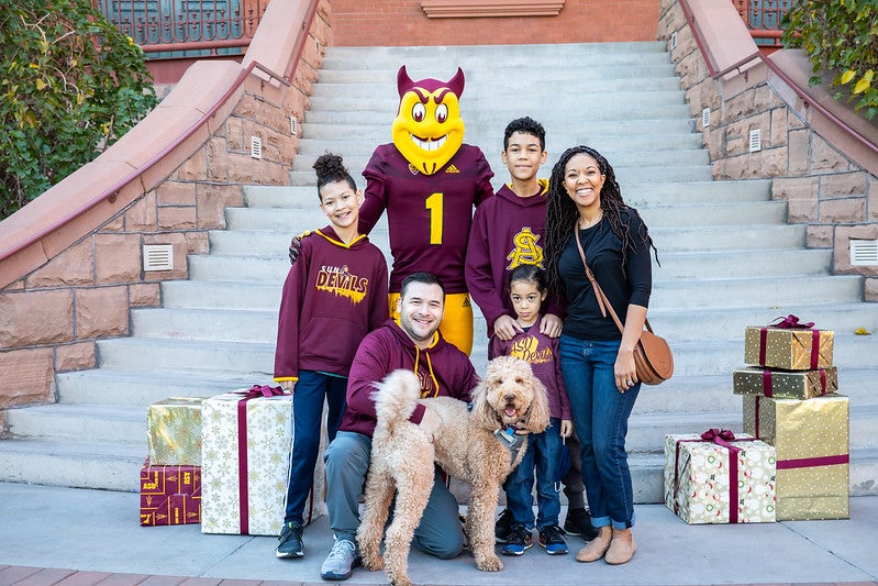 A family of five poses for a family photo with Sparky and their dog in front of Old Main. There are presents arranged around them as props. It is day time. 