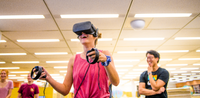 young woman wearing virtual reality goggles motioning with arms as others look on from background