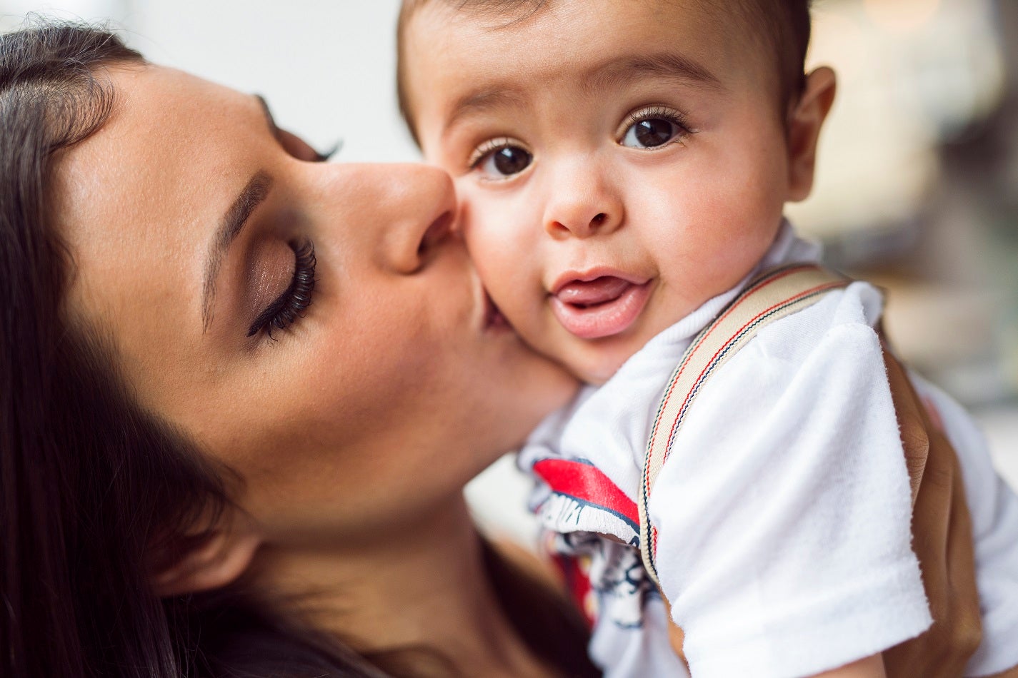 A baby looks out as a woman kisses it on the cheek.