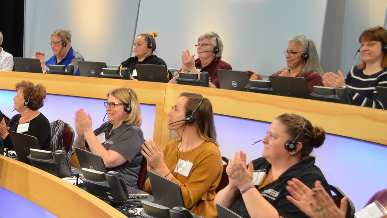 Image of women with phone headsets sitting and clapping behind desks at a pledge drive. 