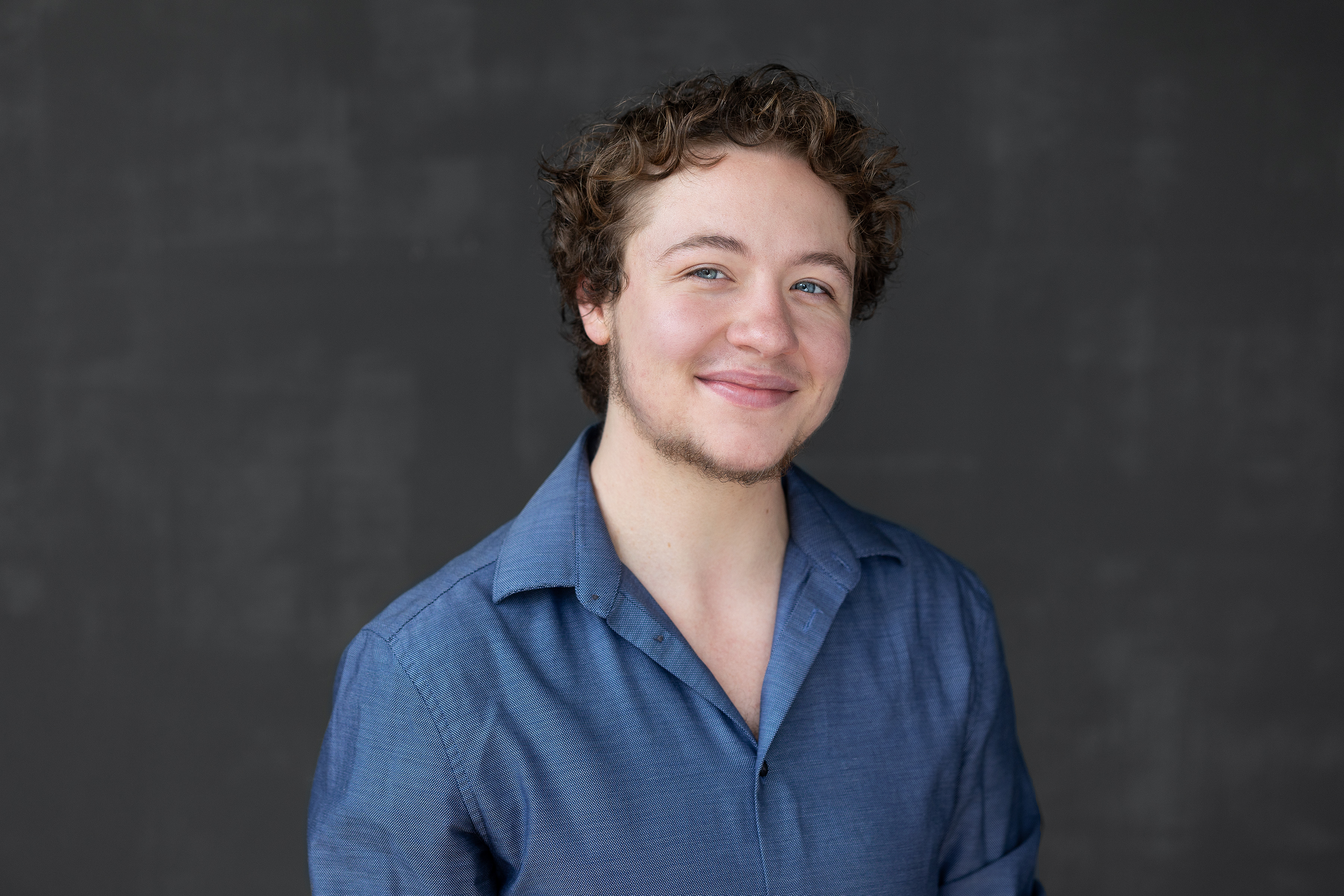 Photograph of Ellis Lane. He has light/white skin and curly brown hair. He's wearing a blue button up shirt and is standing in front of a charcoal background, smiling while looking at the viewer. 